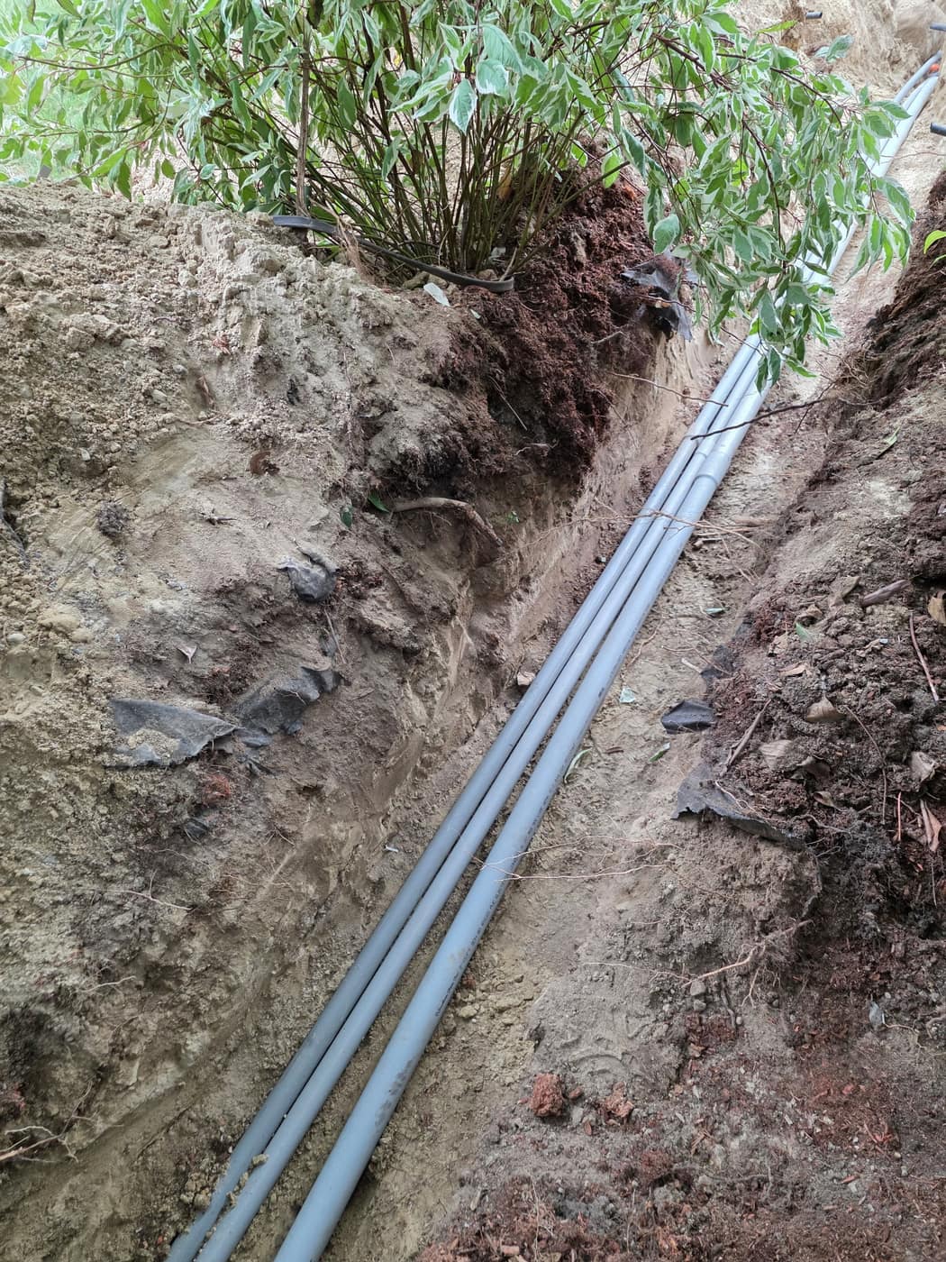 Three grey PVC conduits laid in an open trench beside a landscaped bed