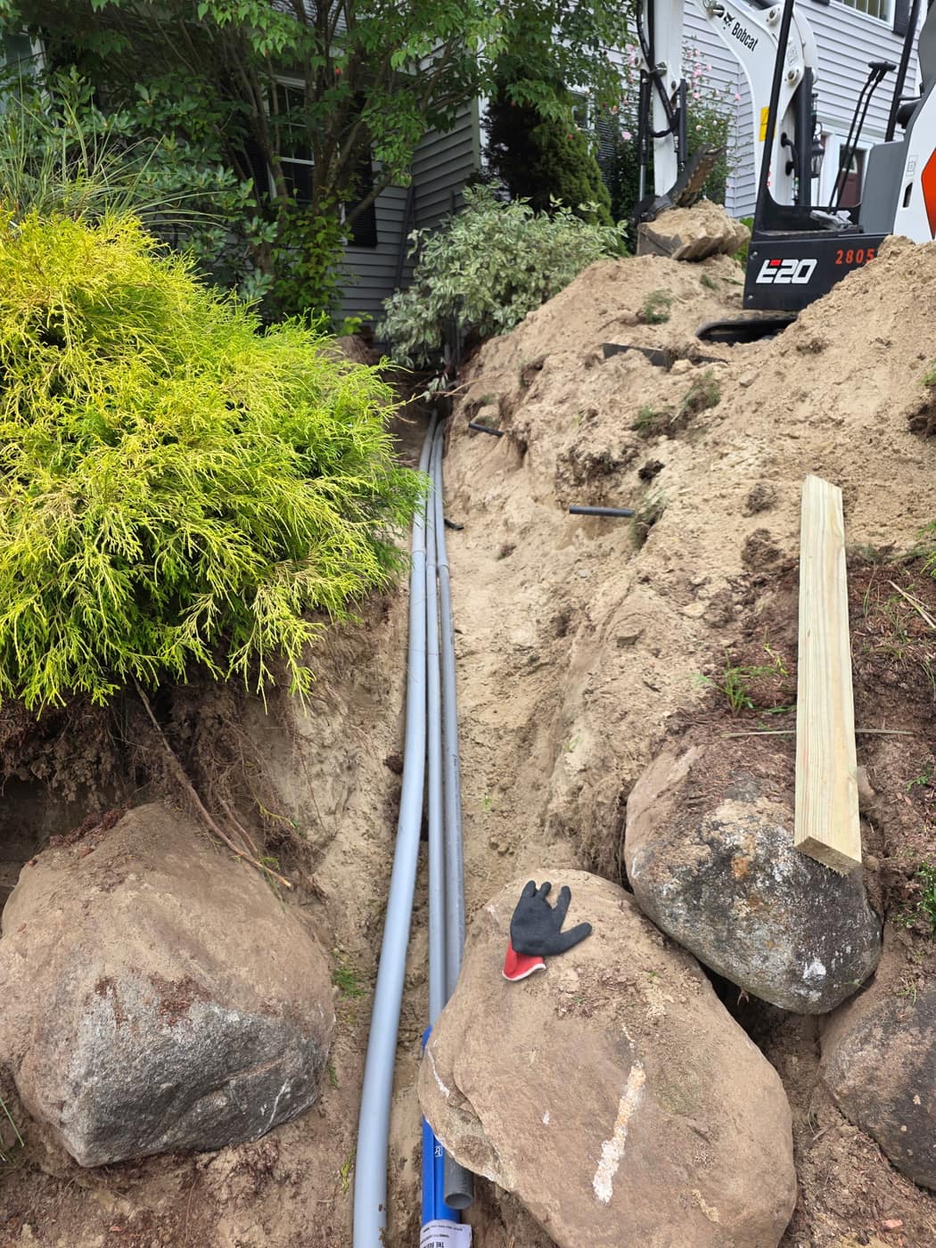 Mini excavator trenching for underground electrical conduit through a landscaped yard with boulders moved aside